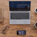 Laptop, Notizheft, Brille und Tasse Kaffee auf Holztisch